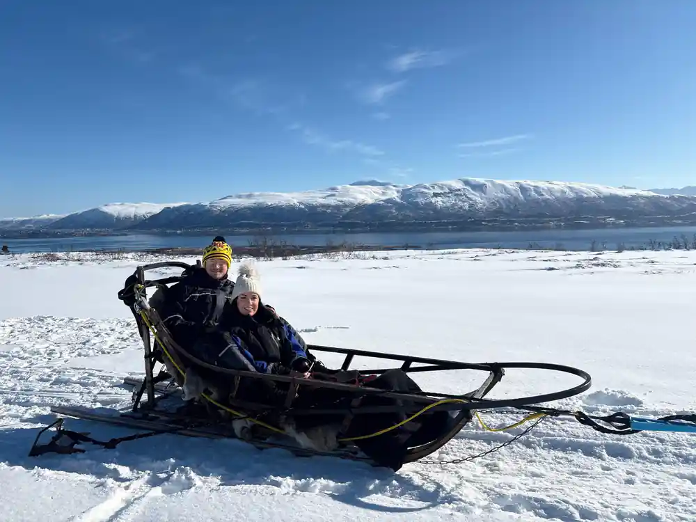Two people sitting in a parked sled on a frozen lake below the mountains
