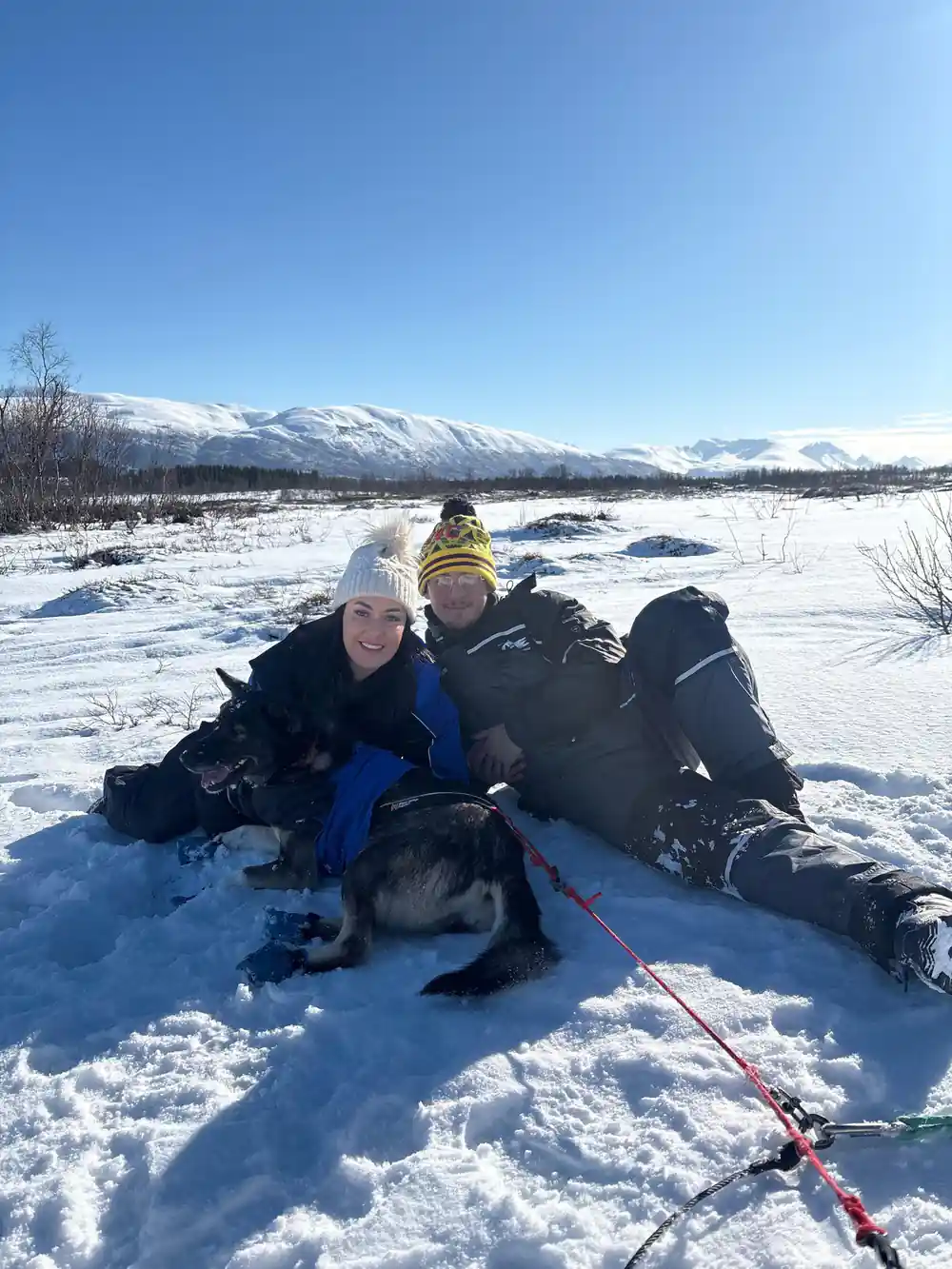 Two visitors lying in the snow with a sled dog between them