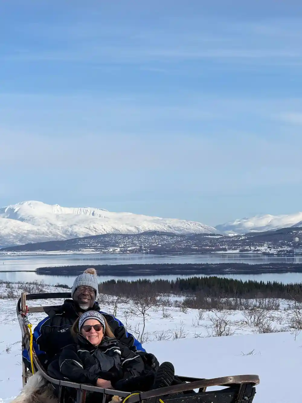 Two visitors in the sled basket with the fjord and peaks behind