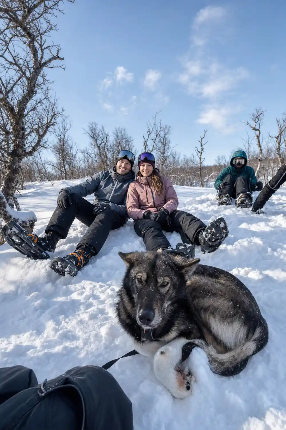 Two visitors in thermal suits sitting in the snow with a dog