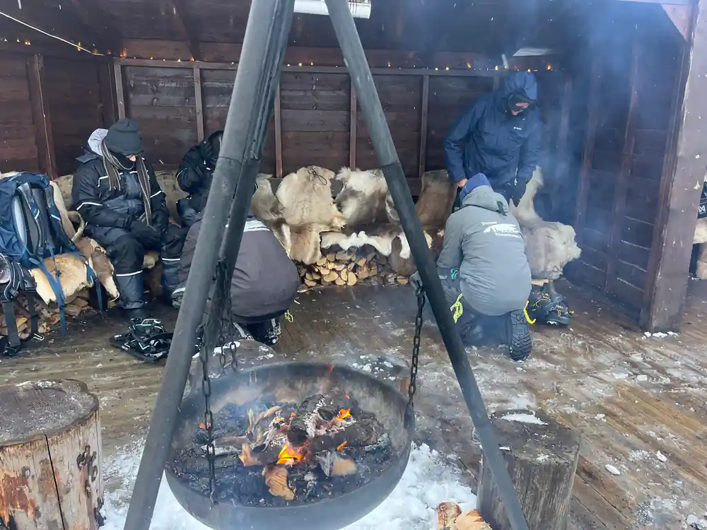 Visitors around an open fire pit under a timber shelter with hides